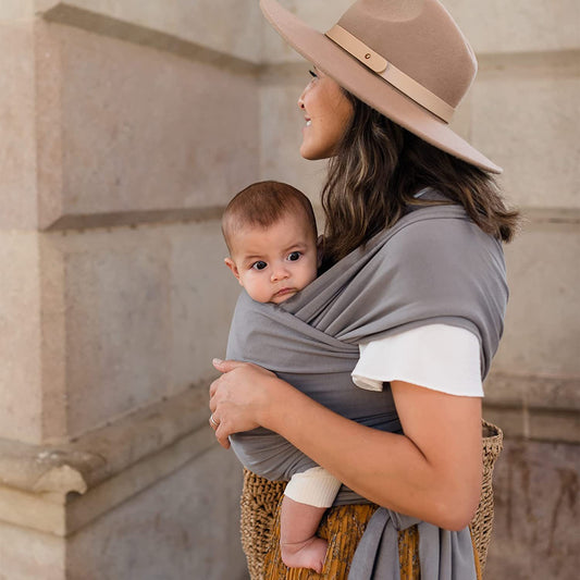 Woman wearing a soft gray ergonomic baby wrap carrier with a baby facing forward, outdoors.