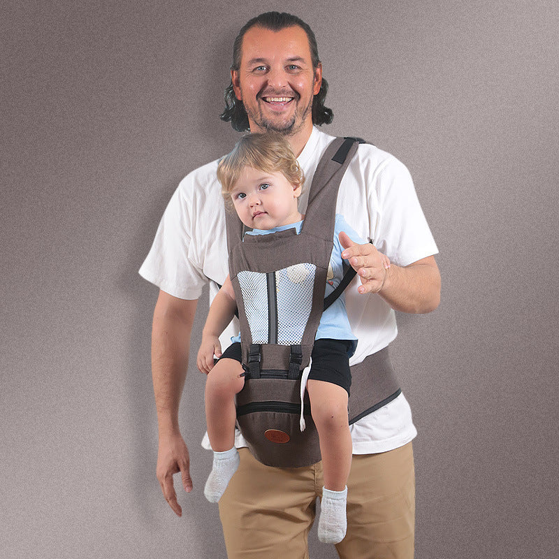 Smiling man carrying a toddler in a brown ergonomic baby hip carrier with breathable mesh.