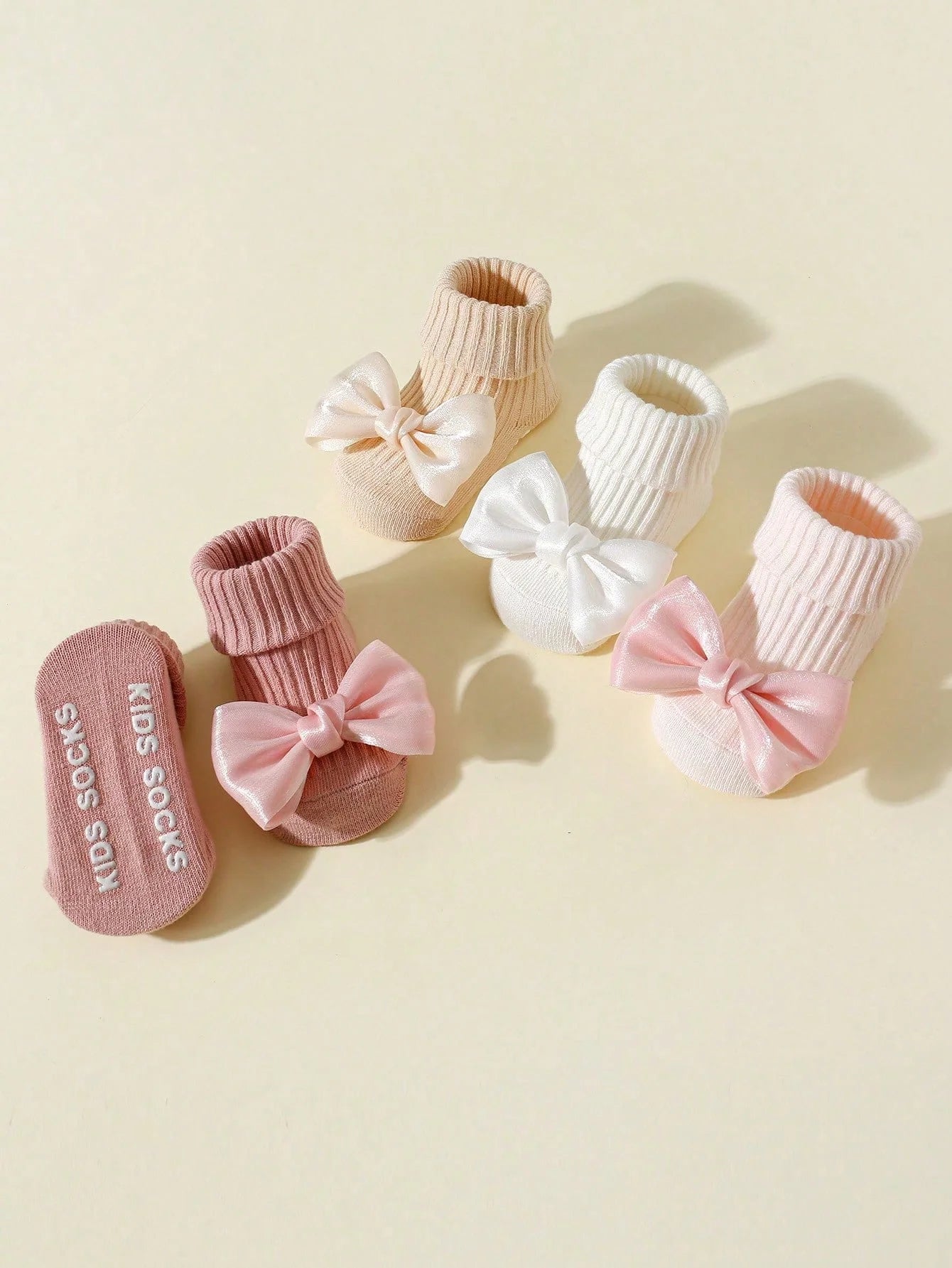 Four pairs of cotton baby socks with bows in pink, white, and beige, featuring non-slip soles.