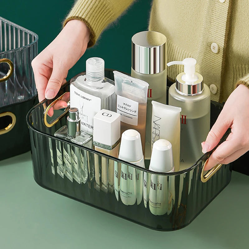 Clear rectangular storage box with gold handles, holding cosmetics and skincare products.