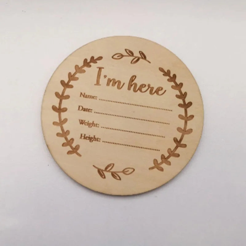 Round wooden birth announcement sign with "I'm here" and fill-in blanks for name, date, weight, and height, framed by engraved leaves.