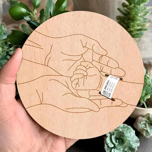 Round wooden birth keepsake with engraved hands holding a hospital bracelet, surrounded by green plants.
