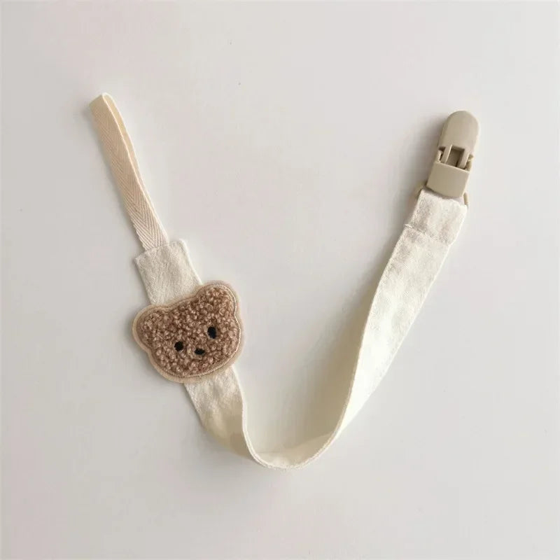 Beige cotton pacifier clip with a cute embroidered brown teddy bear design and a plastic clasp.