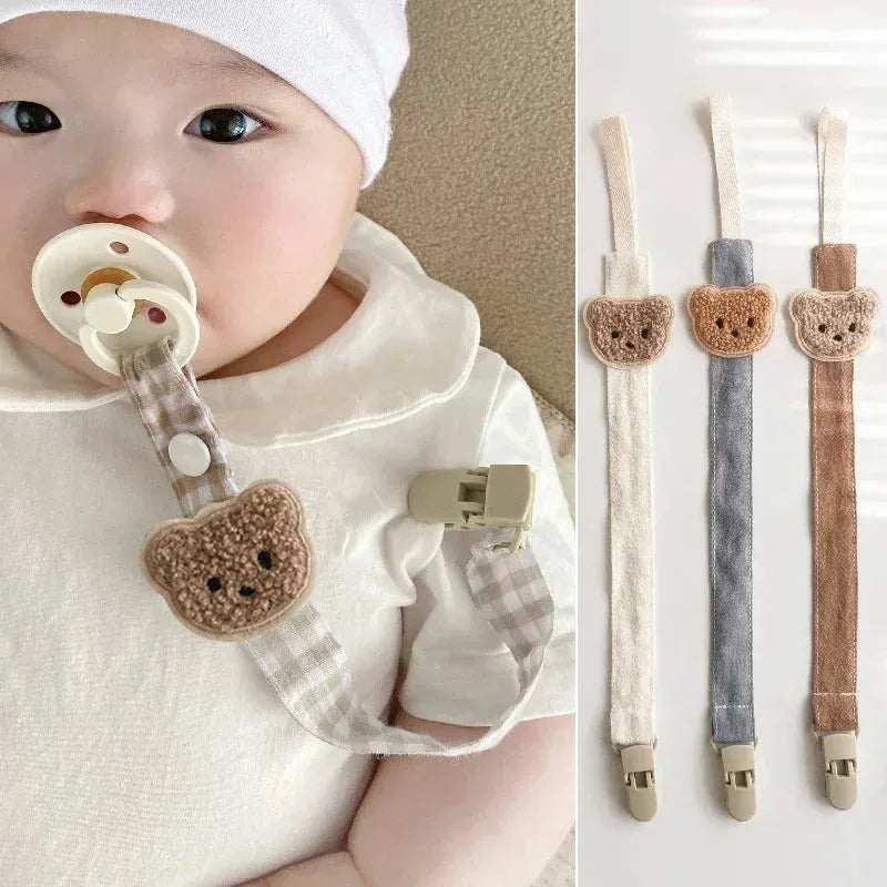 Baby with a beige pacifier clip featuring a brown embroidered bear and checkered cotton strap. Three clips in white, gray, and brown are shown.