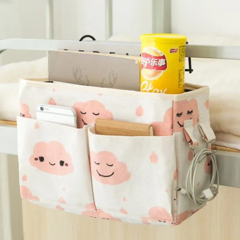 Pink and white cotton multi-pocket bedside organizer with cute cloud print, holding a phone, book, and snacks.