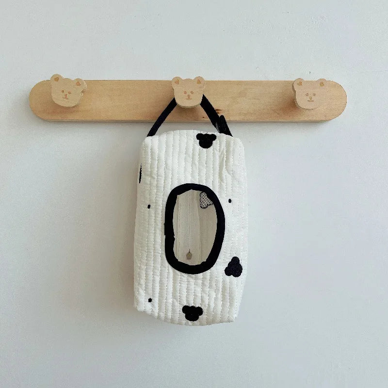 White quilted cotton hanging tissue and wipe dispenser with black spots and oval opening, on a wooden hook.