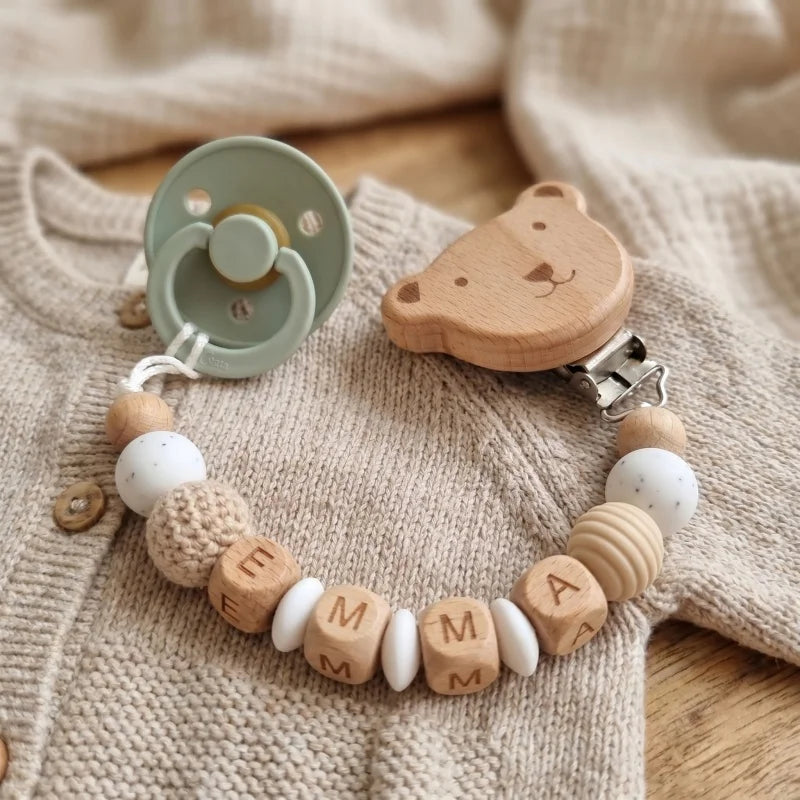 Personalized wooden and silicone pacifier clip with "EMMA" beads and a bear-shaped clip, next to a sage green pacifier on a beige knitted outfit.