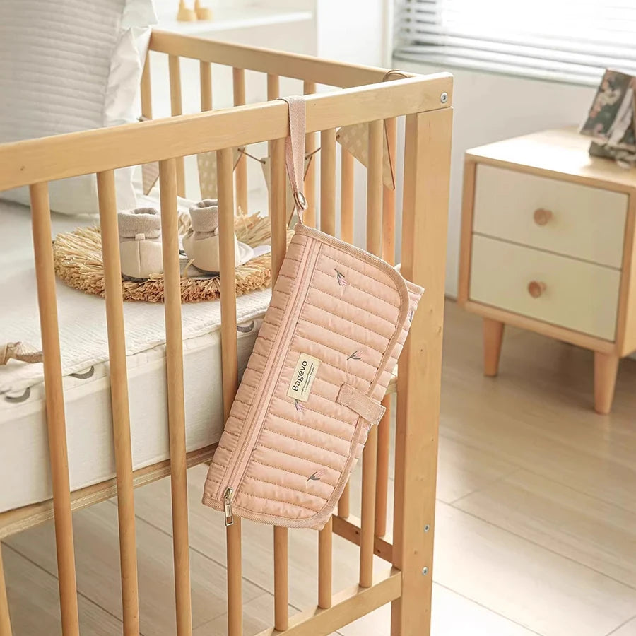 Pink quilted portable changing pad with storage pouch hanging on a wooden crib.