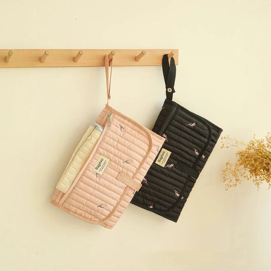 Two quilted, portable baby changing pads, one pink with a leaf pattern and one black, hanging on a wooden rack.
