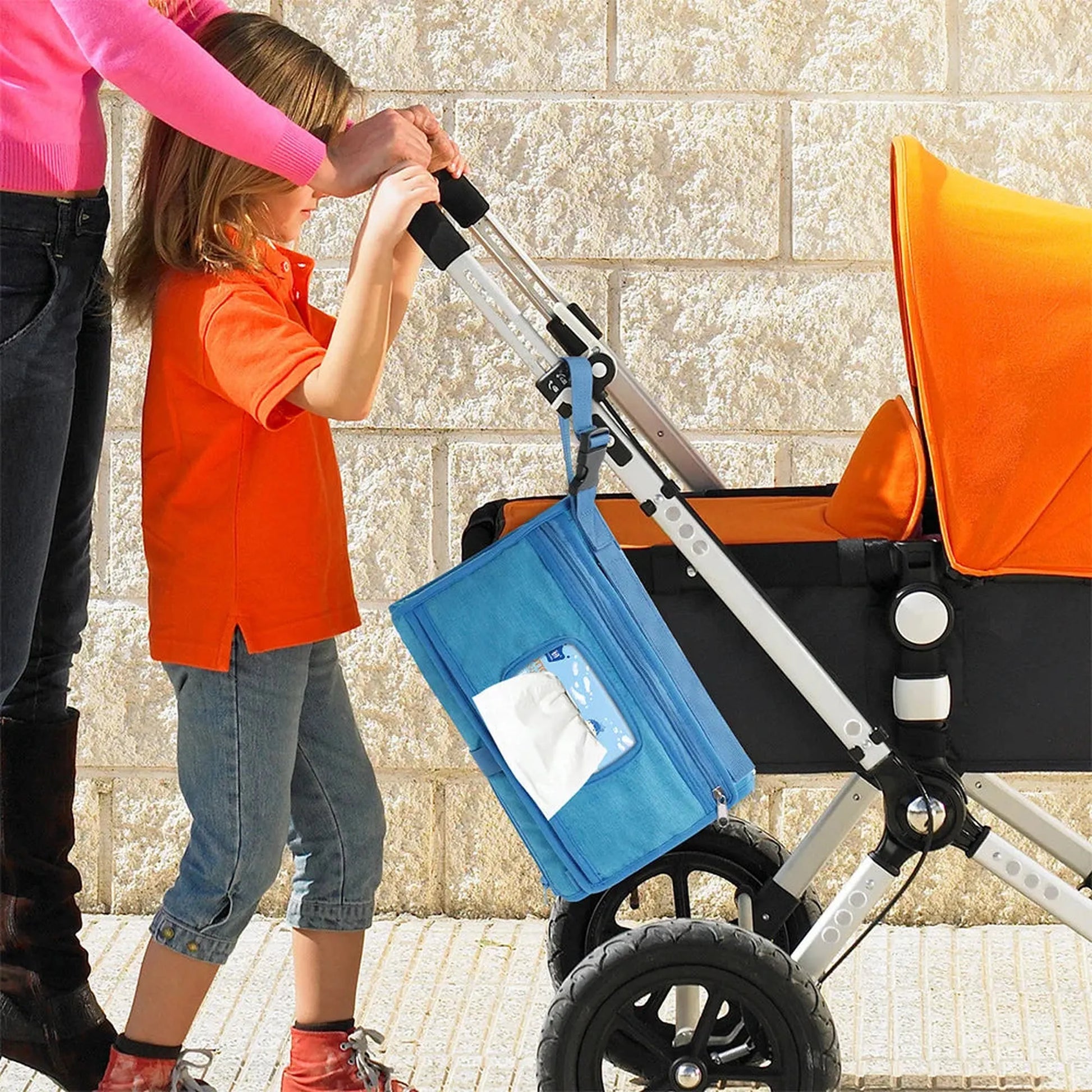 Portable blue changing pad with clear pocket attached to a stroller, next to an adult and child.