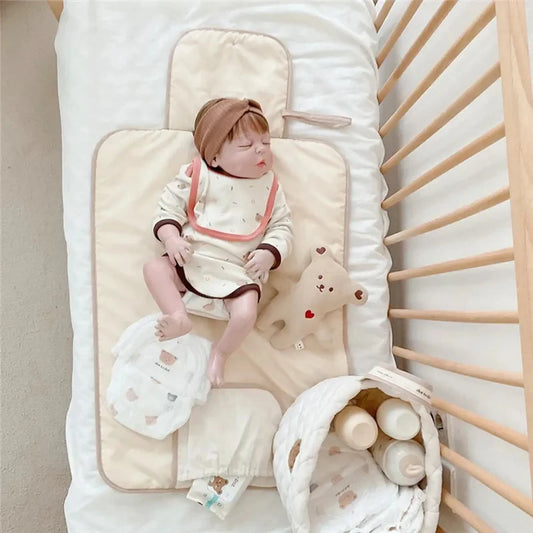 Portable, foldable, waterproof cotton changing mat in beige with a sleeping baby, diapers, and a bear toy.