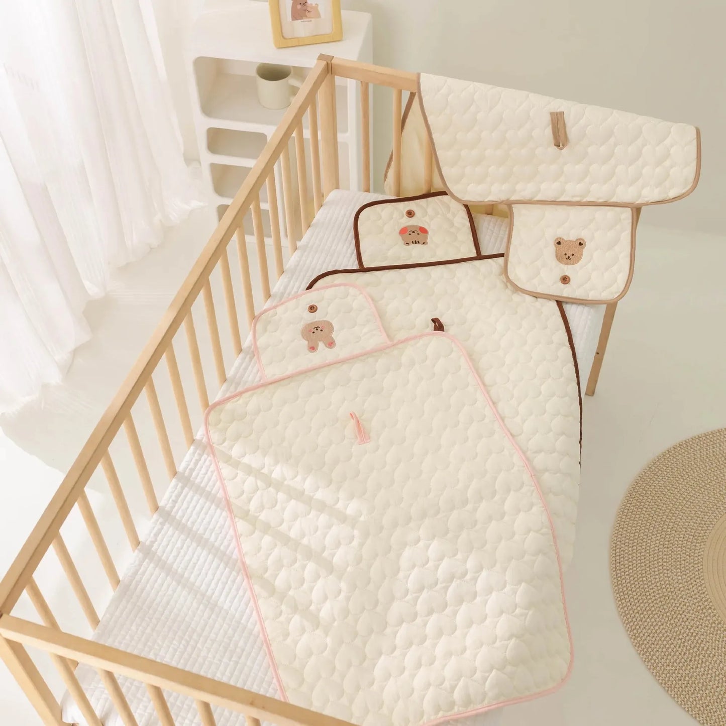 Portable, quilted cream cotton changing pad with brown trim and bear embroidery, shown folded and unfolded in a crib.