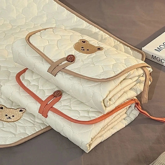 Foldable, portable cream cotton changing pad with quilted texture, brown trim, and embroidered bear, secured with a button.