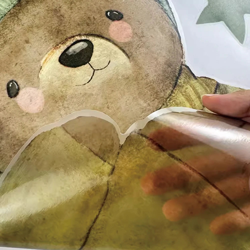 Hand peeling a brown bear wall decal with pink cheeks and green accents from its backing.
