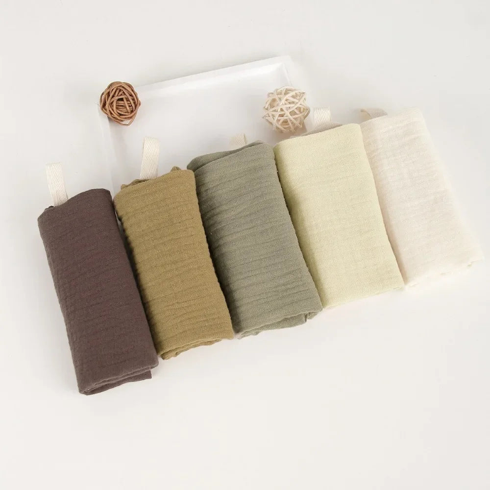 Five folded baby gauze cotton towels in brown, olive, sage, cream, and off-white, with hanging loops.