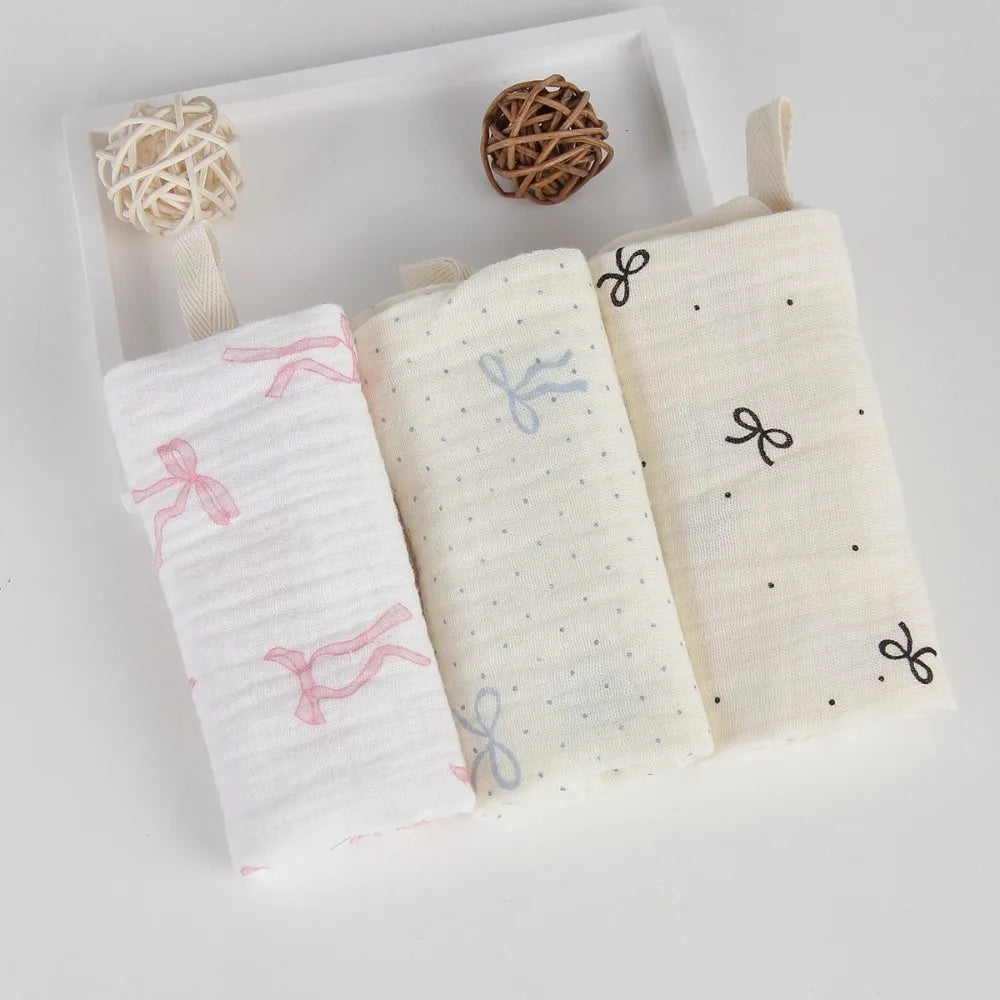 Three soft white cotton gauze baby towels with bow patterns, folded on a tray.