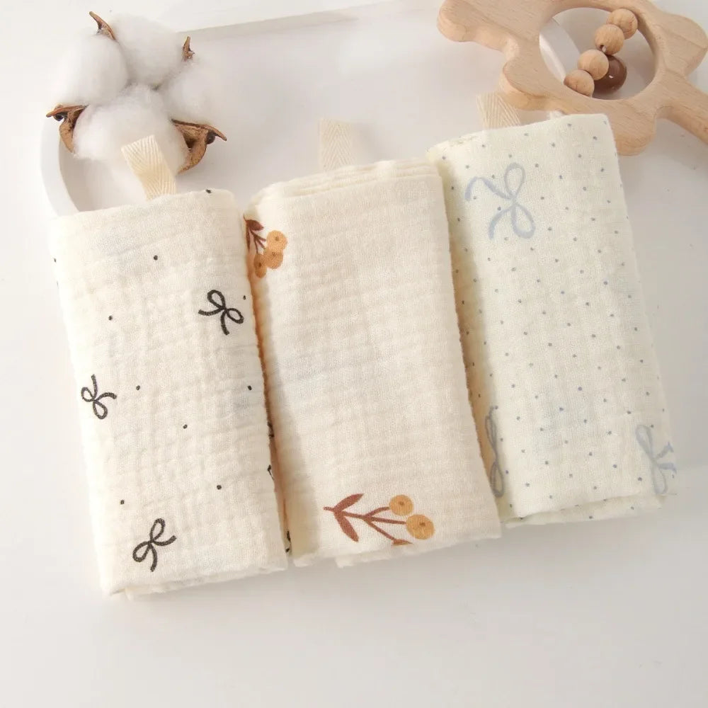 Three soft, natural cotton gauze baby towels, off-white with delicate bow, floral, and dot patterns.