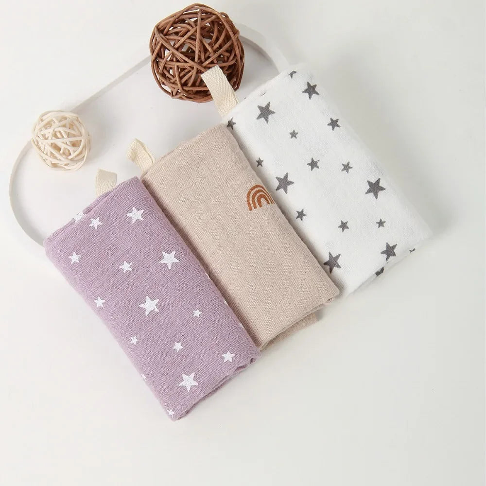 Three soft cotton gauze baby towels: purple with white stars, beige with a rainbow, and white with gray stars.