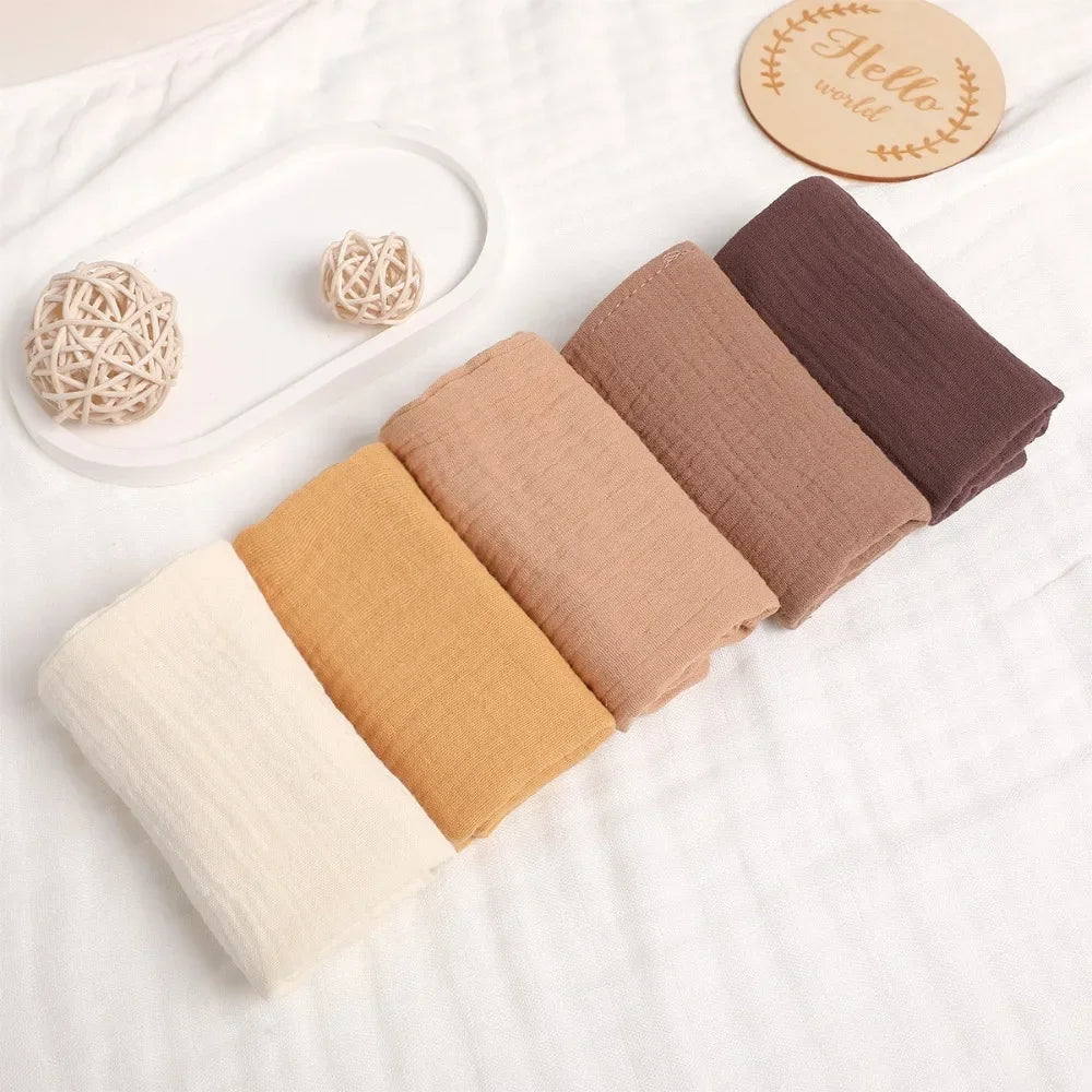 Soft multi-layered gauze cotton baby towels in a natural earth-tone palette, including cream, mustard, and brown shades.