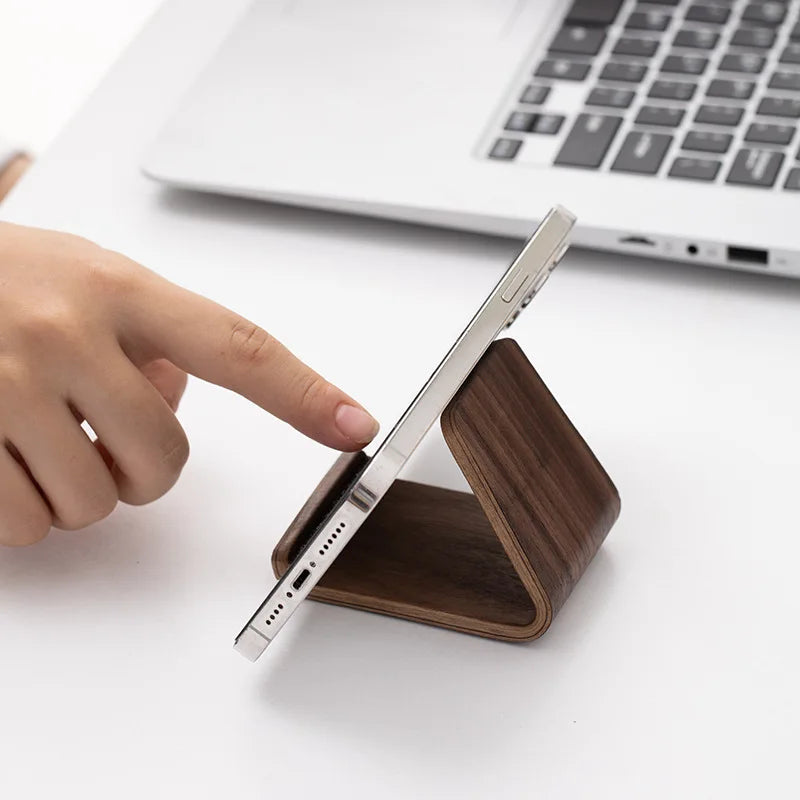 Minimalist dark walnut wood phone stand holding a silver smartphone on a white desk, with a hand touching the screen.