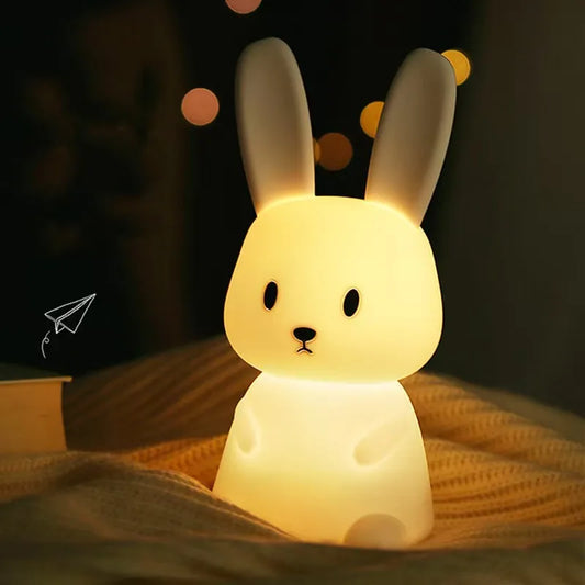 Silicone rabbit night light with touch sensor, glowing warm yellow on a textured blanket.