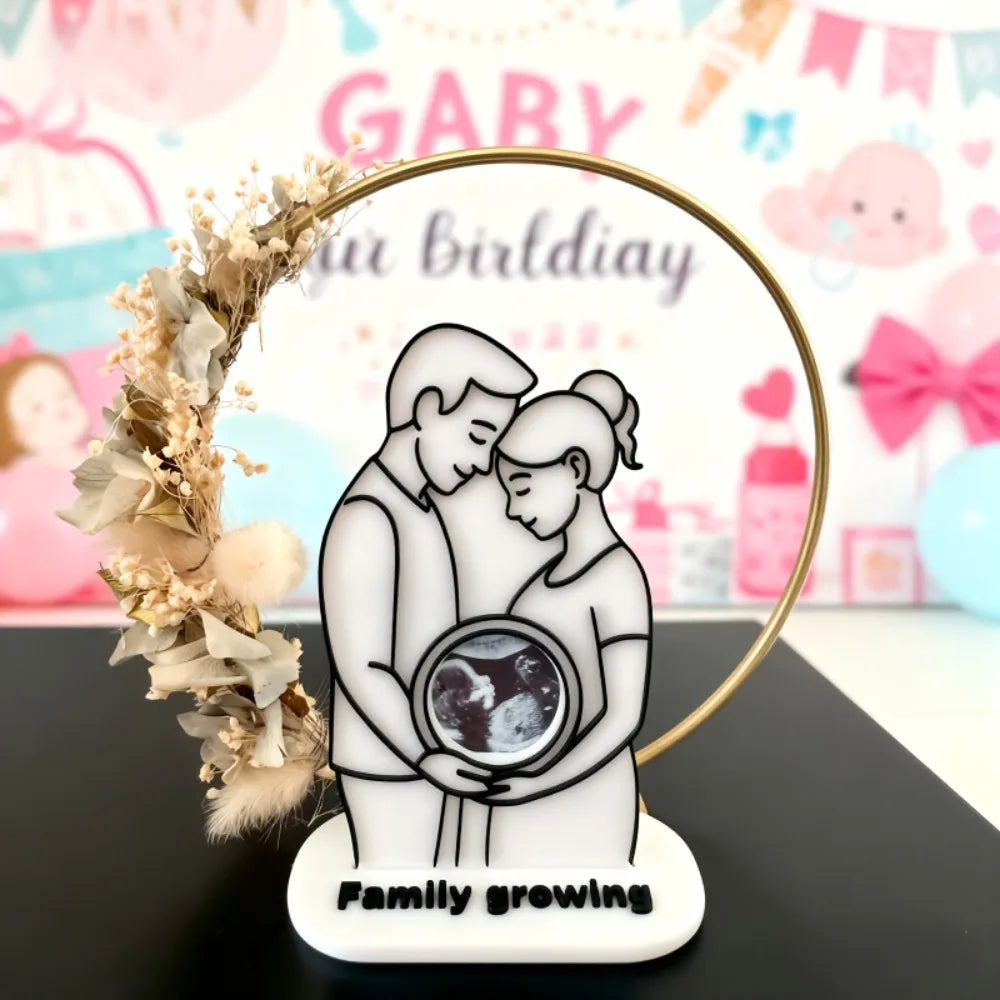 White plastic ultrasound photo frame with a couple holding a baby scan, adorned with a gold ring and dried flowers.