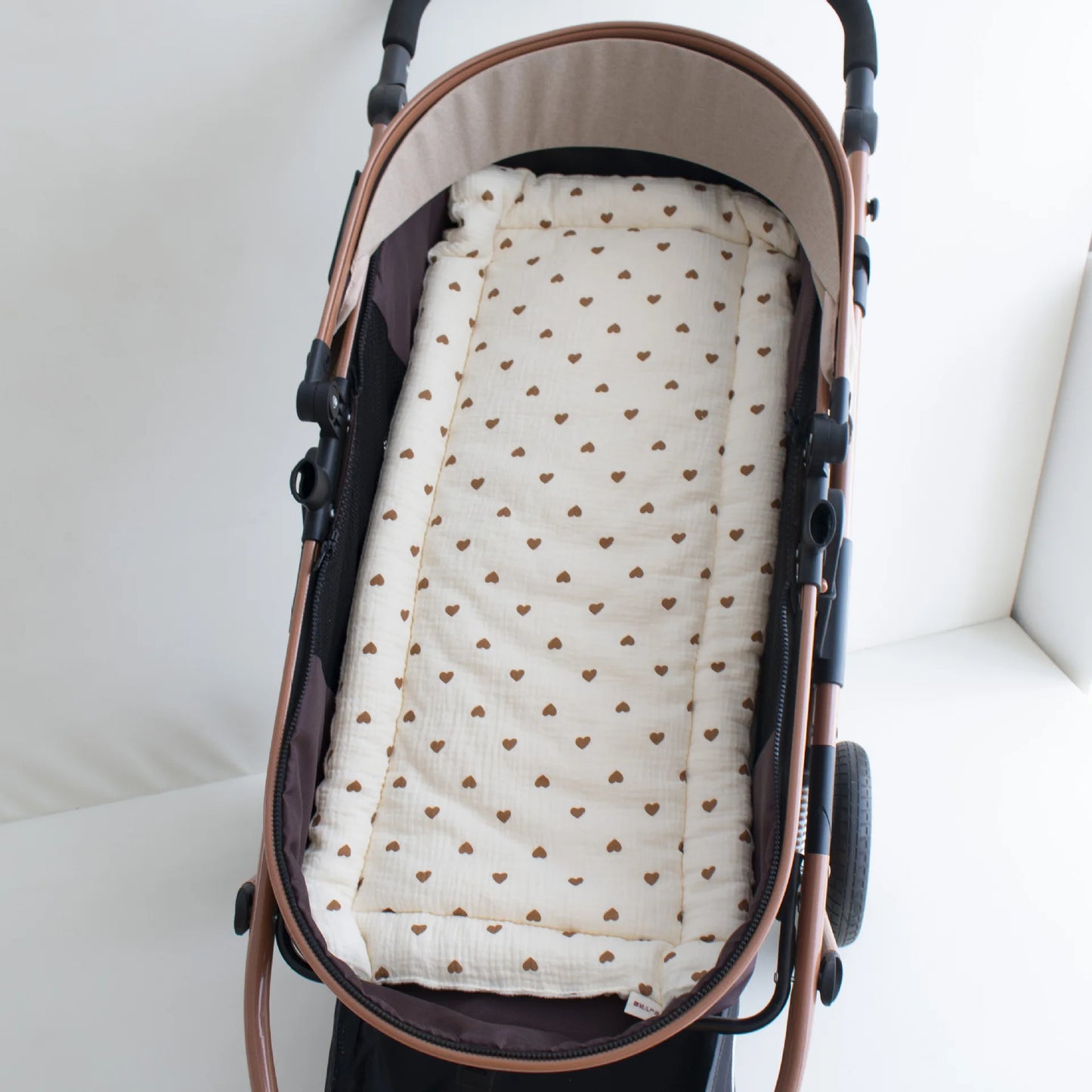 Soft, cream-colored cotton stroller cushion with brown heart pattern, fitted in a rose gold stroller.