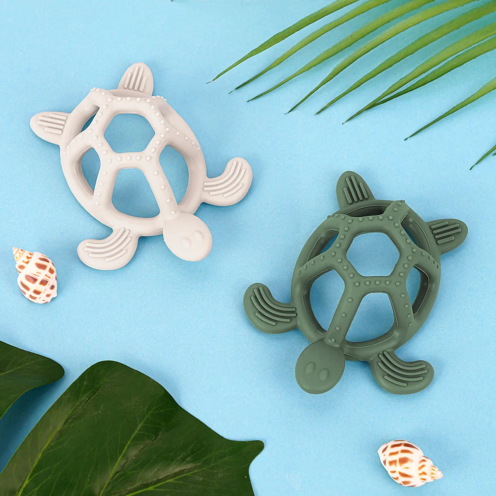 Two turtle-shaped silicone baby teethers, one light gray and one dark green, on a blue background with leaves and seashells.