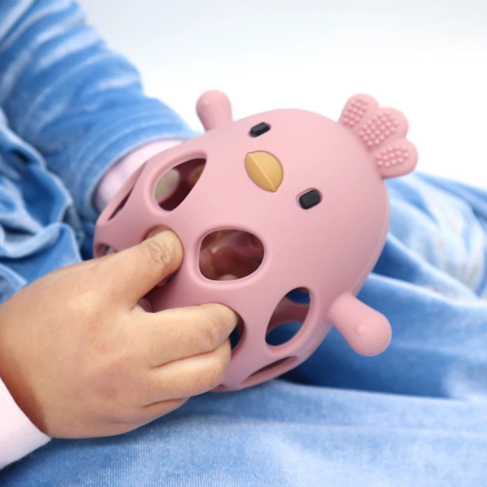 Baby's hand holding a pink, hollow silicone chick teether with a textured comb.