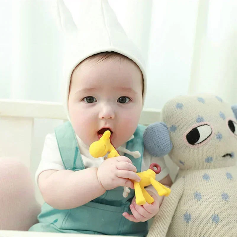 Baby in a white hat and teal overalls chewing on a yellow giraffe silicone teether.