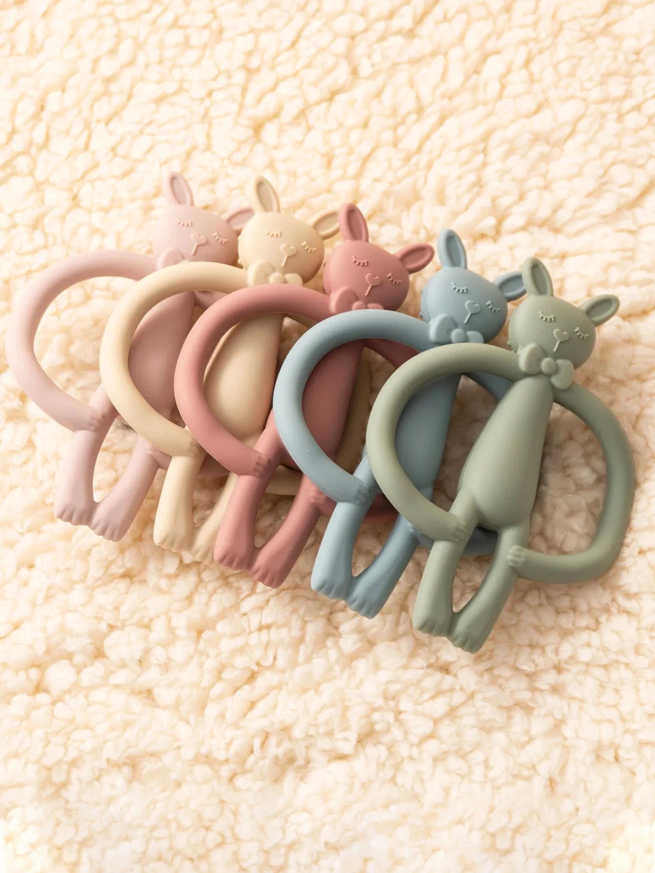 Soft silicone bunny teethers in pastel pink, yellow, peach, blue, and green on a cream fuzzy blanket.