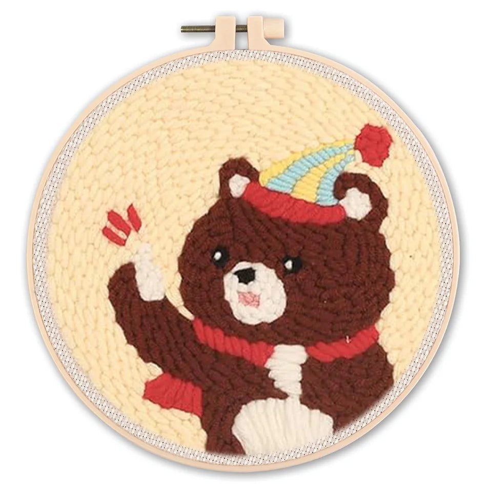 Punch needle embroidery kit featuring a cute brown bear with a party hat and red scarf, in a round hoop.