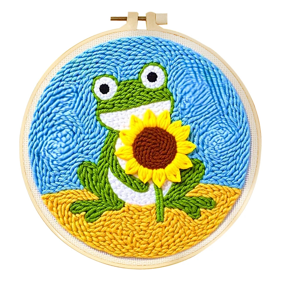 Punch needle embroidery kit featuring a green frog holding a yellow sunflower against a blue sky, framed in a wooden hoop.