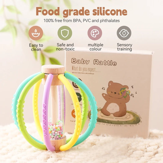 Wooden baby rattle with colorful silicone rings, rain drum, and teether, next to its packaging.