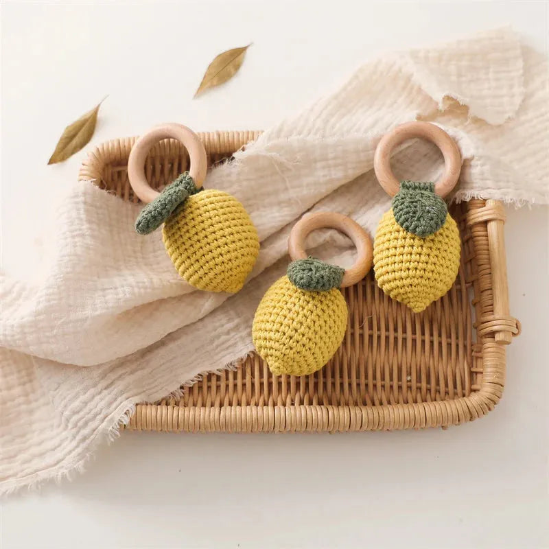 Three yellow crocheted lemon rattles with natural wood rings on a wicker tray with a white muslin cloth.