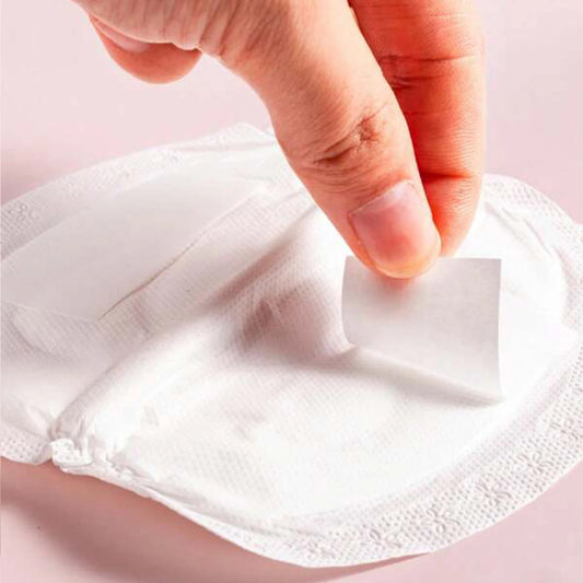 Hand peeling the adhesive backing from a white, thin, disposable nursing pad on a pink surface.