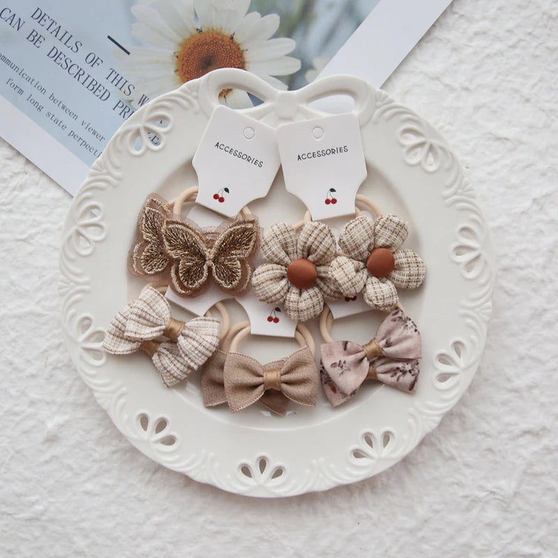 Set of 6 girls' elastic hair ties with floral, butterfly, and bow designs in neutral fabric.