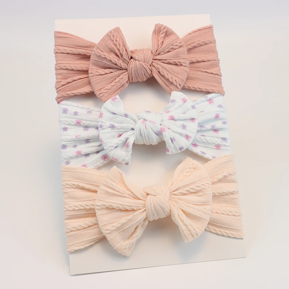 Set of 3 soft nylon baby headbands: pink, floral white, and peach with bow designs.