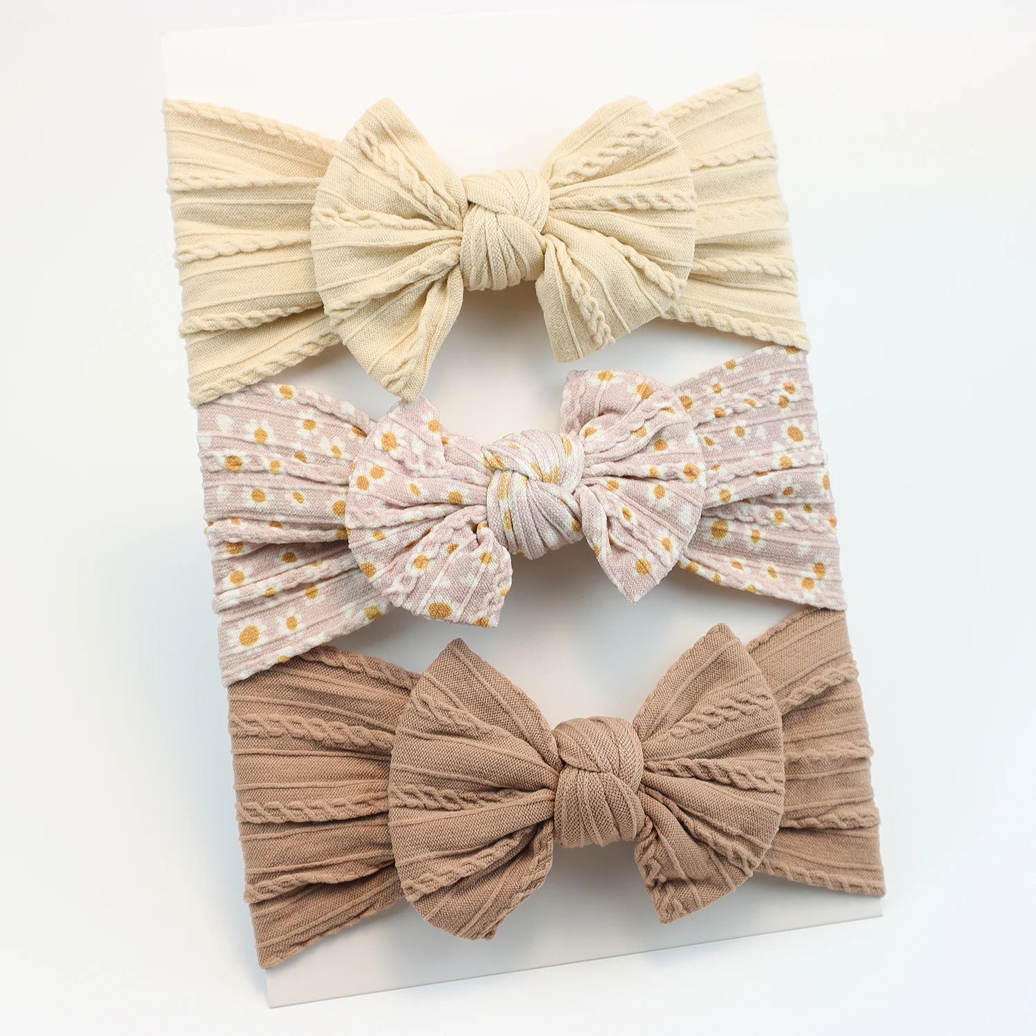Set of 3 soft nylon baby headbands: cream, floral pink, and brown, each with a decorative bow.