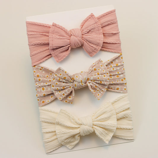 Set of 3 soft nylon baby headbands: pink ribbed, floral print, and cream cable knit bows.