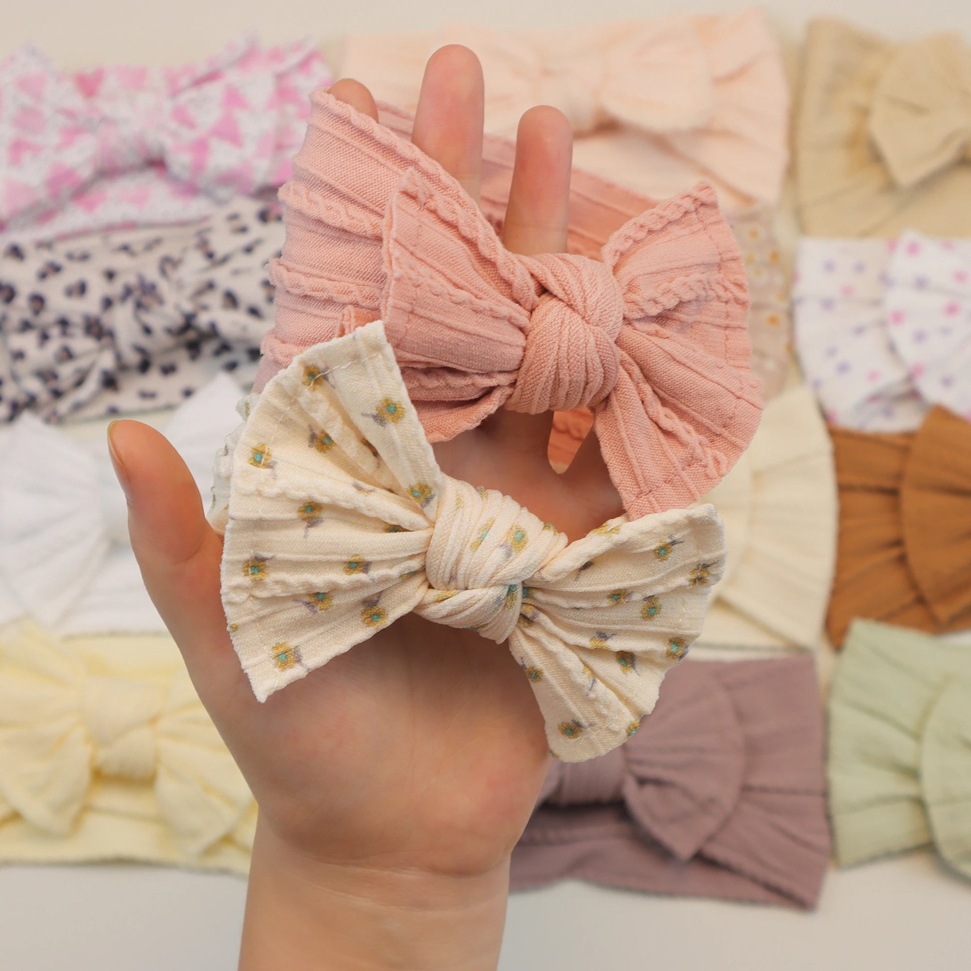 Hand holding two soft, flexible nylon baby headbands: one ribbed pink, one cream with tiny floral print.
