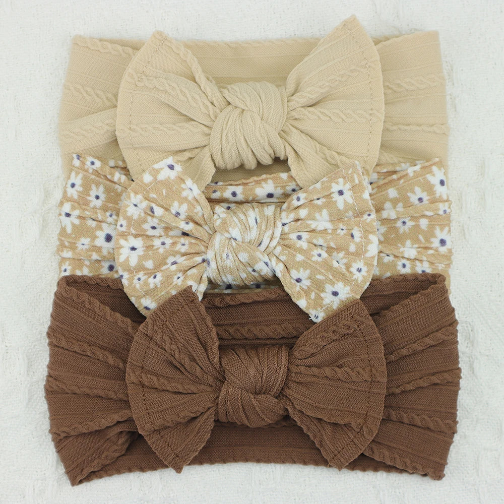Set of 3 soft nylon baby headbands: beige cable knit, floral print, and brown ribbed with bows.