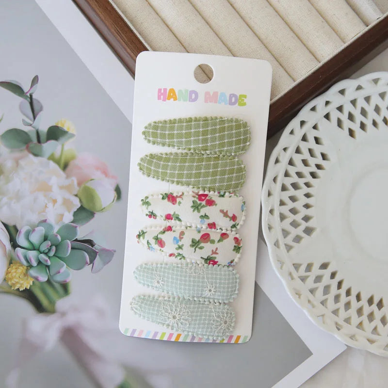Six handmade fabric snap clips for girls, featuring green gingham, floral, and light blue lace designs.