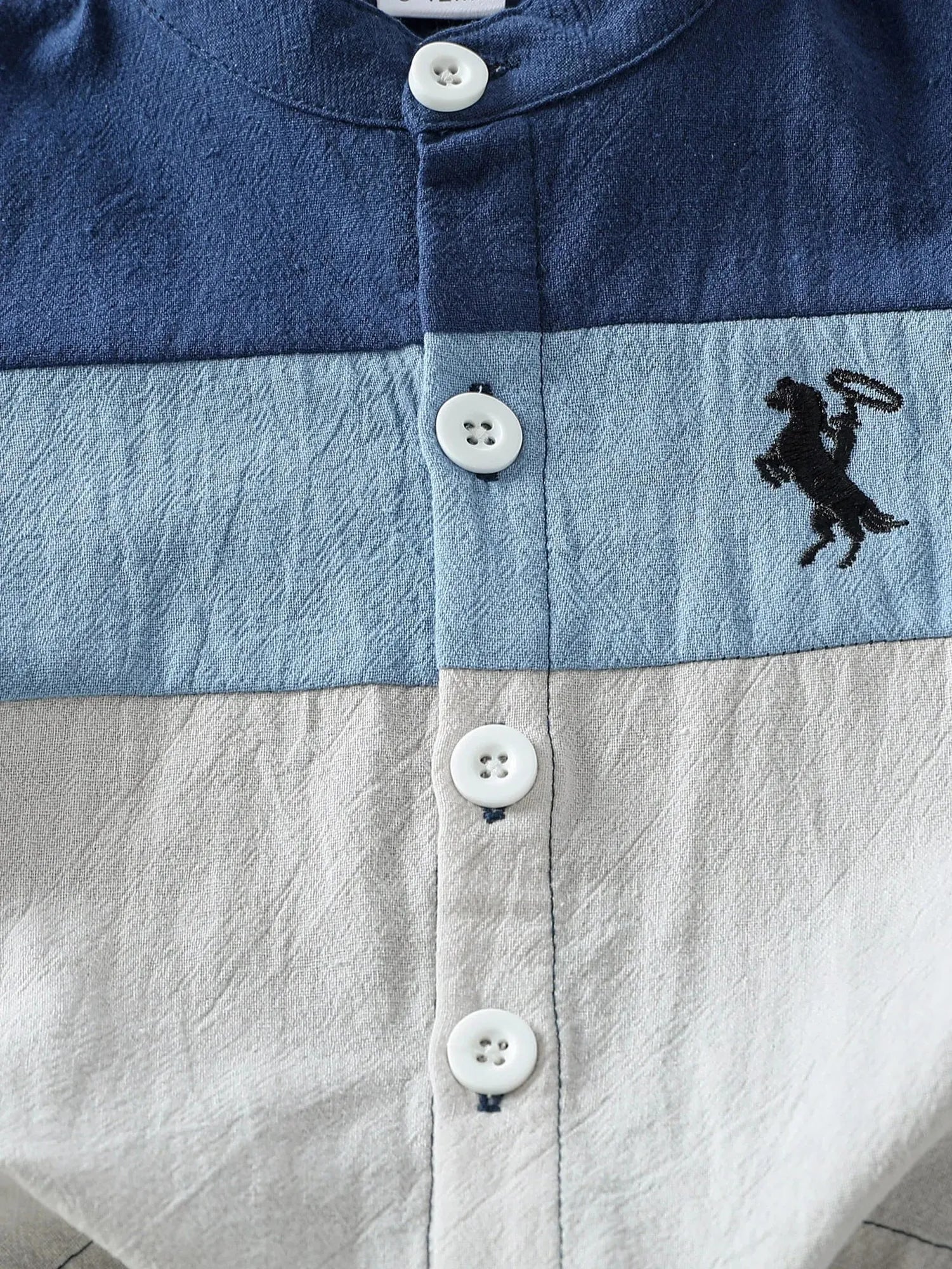 Close-up of a baby boy's short-sleeve cotton summer shirt with blue, light blue, and grey color blocks and a horse embroidery.