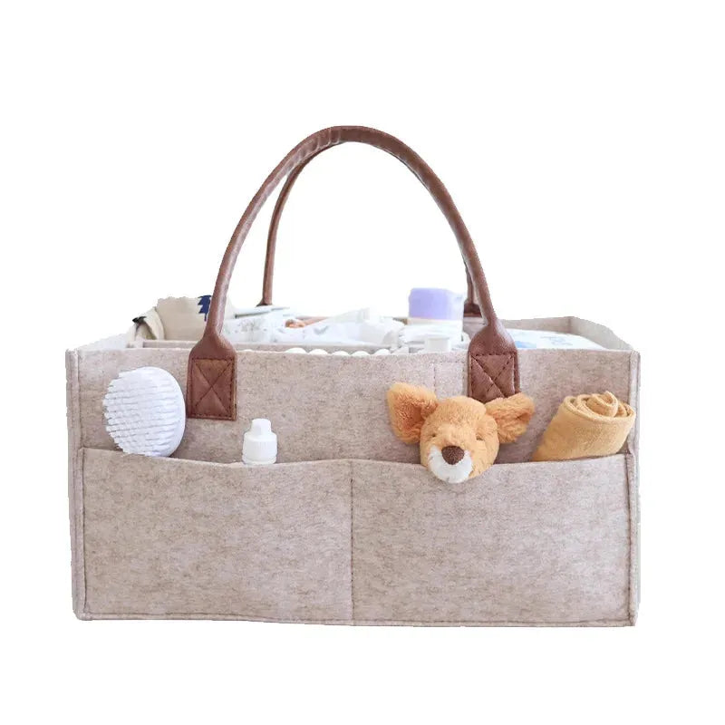 Beige felt diaper caddy with brown faux leather handles, filled with baby essentials and a plush toy.