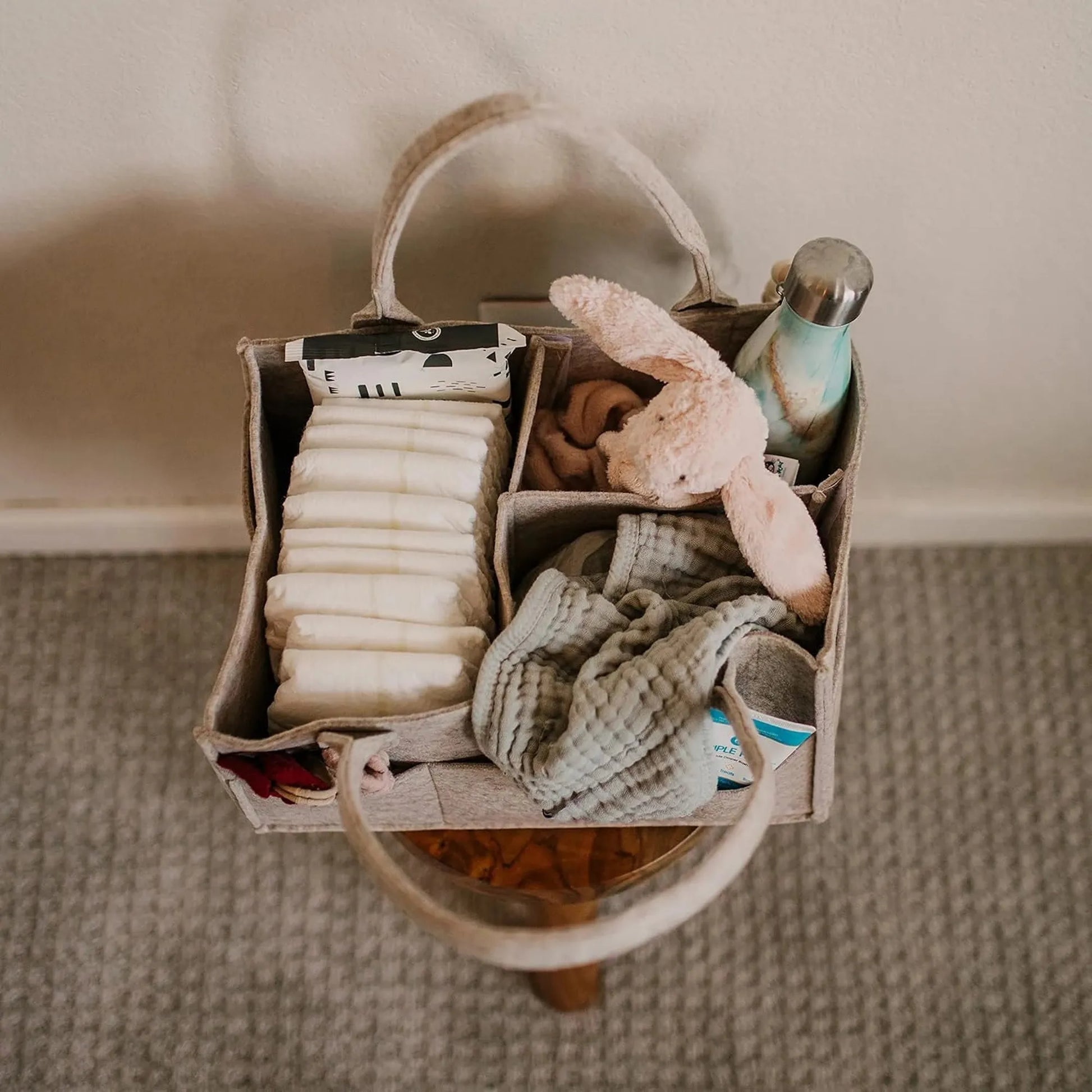 Beige felt baby diaper caddy with handles, filled with diapers, a plush bunny, clothes, and a water bottle.