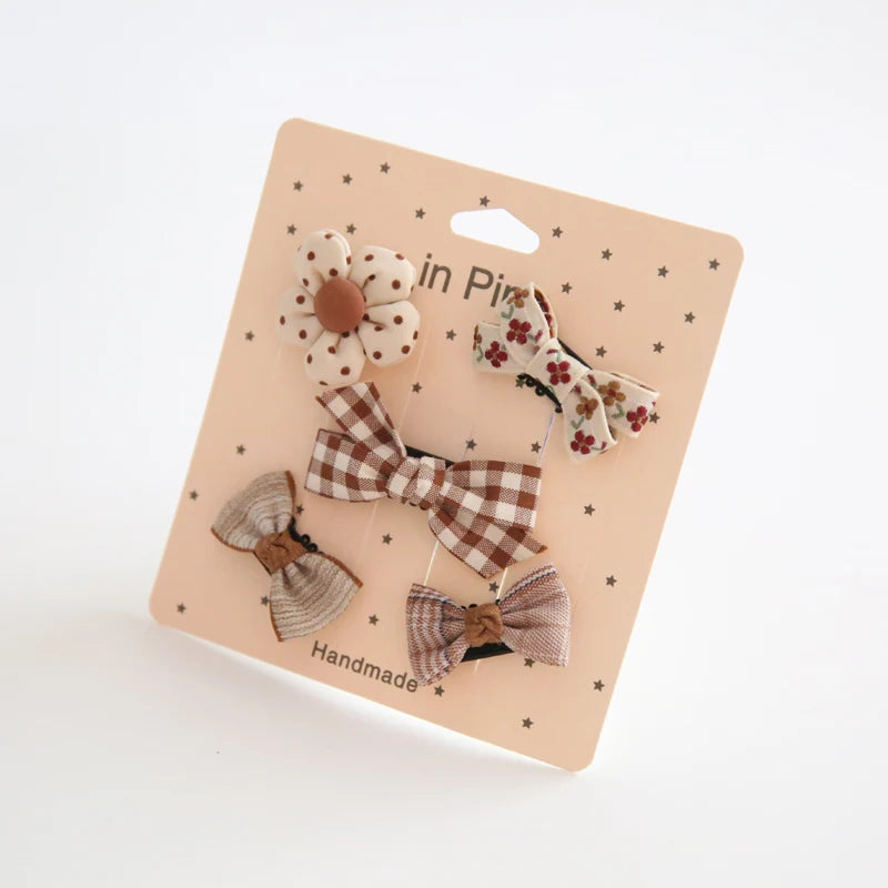 Set of five cute baby hair clips: a flower, two plaid bows, and two patterned fabric bows, on a pink card.