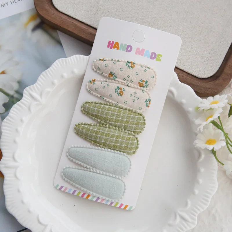 Handmade set of 6 fabric snap hair clips for babies and girls, featuring floral, plaid, and solid designs in pastel green and cream.