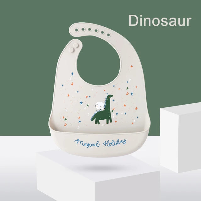 Adjustable silicone baby bib in light beige with a green dinosaur print and "Magical Holiday" text.