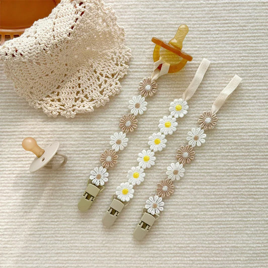 Three daisy flower pacifier clips in white, yellow, and beige, with a pacifier and crochet bib.